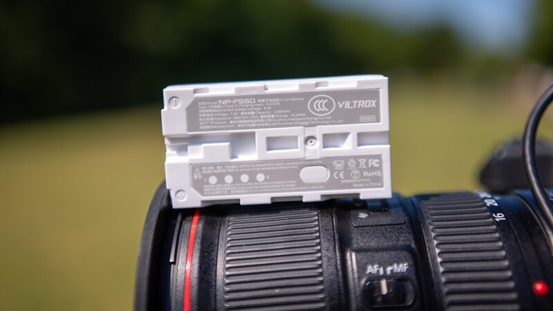 A Viltrox battery grip mounted on top of a telephoto lens with a blurred outdoor background.