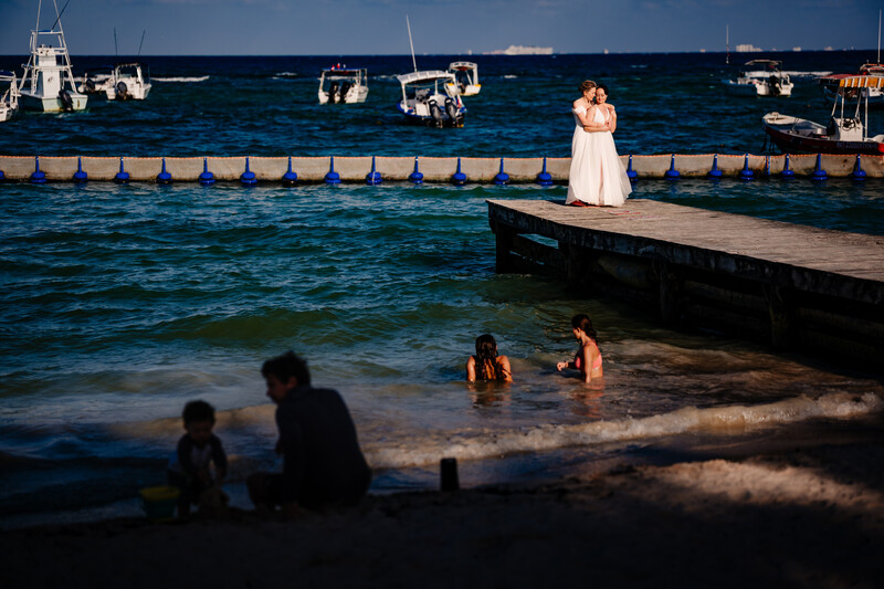 Bride in white dress standing on wooden dock above shallow water with boats and beachgoers in foreground.