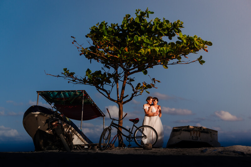 Couple in white wedding attire standing beside a bicycle under a windswept tree at sunset.