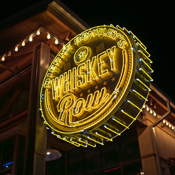Vintage neon sign for Whiskey Road glowing golden yellow against dark night sky and wooden building facade.