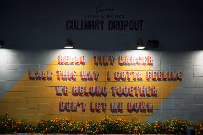 Neon sign with red lettering on yellow brick wall reads "Hello, may I offer check this way I gotta feeling we belong together don't let me down."