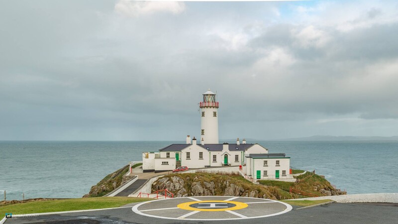 Coastal lighthouse on rocky island with white keeper's house and yellow helicopter landing pad in foreground.