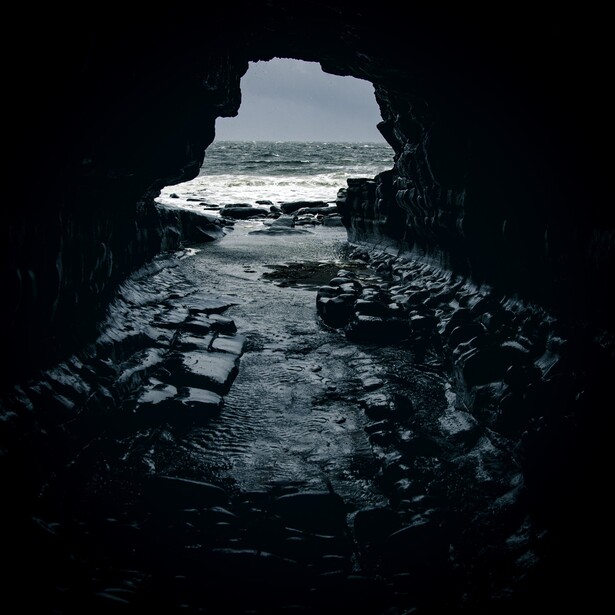 View through a dark rocky cave opening onto a distant coastal landscape with water and sky.