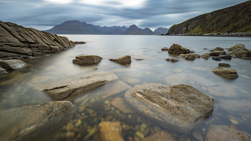 Rocky coastline with submerged boulders in shallow water, mountains across the bay under cloudy sky.