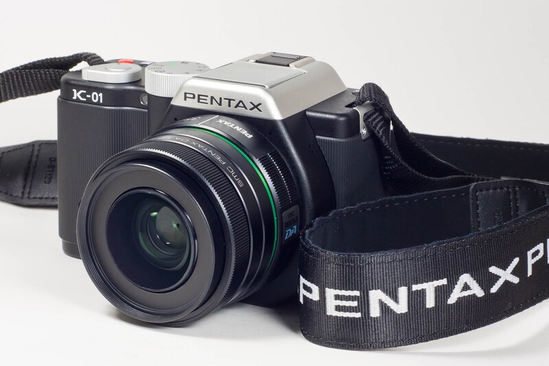 Pentax K-01 digital camera with black lens and shoulder strap against white background.