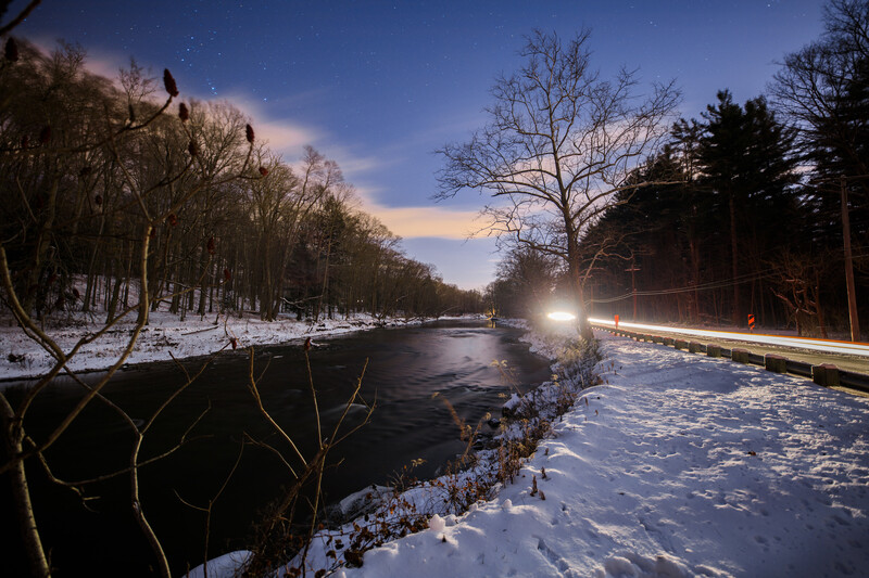 Winter landscape with partially frozen river at dusk, bare trees lining both banks, and car headlights streaking along a road on the right.