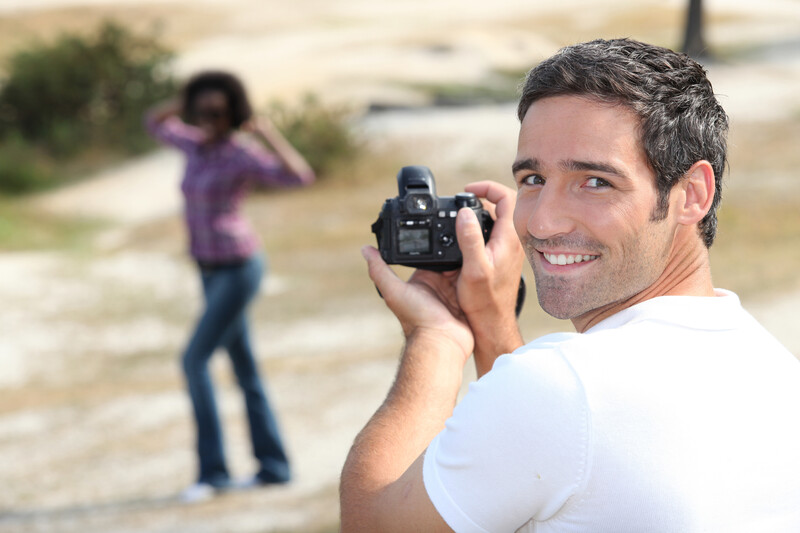 Male photographer smiling at camera while holding a DSLR, with female subject posing in background on rural road.