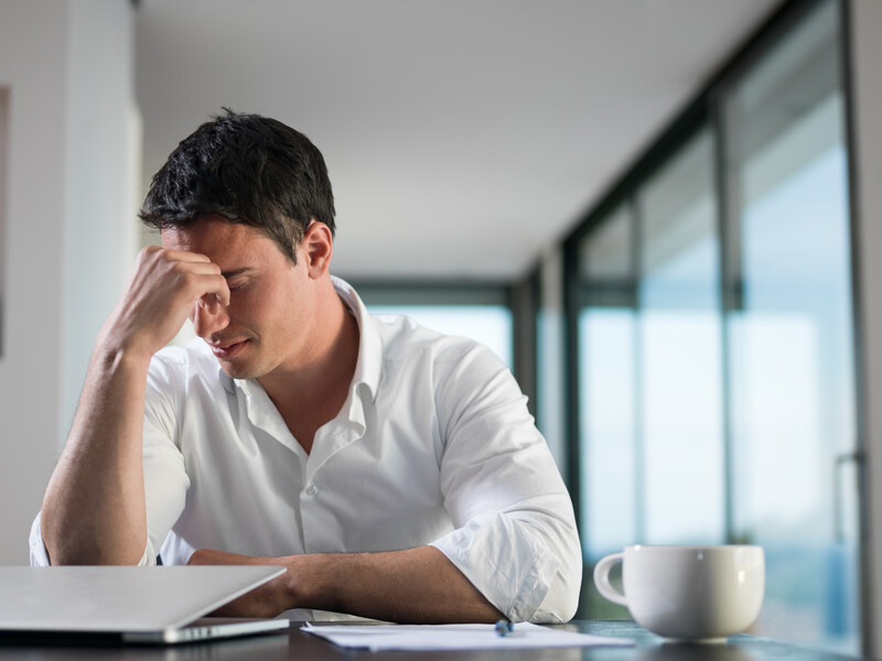 Man in white shirt resting head on hand at desk with laptop and coffee cup.