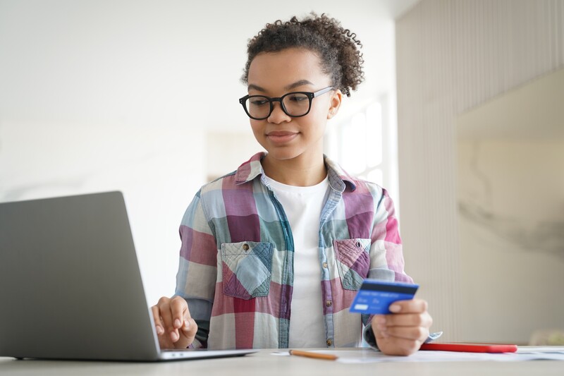 Young person wearing glasses and plaid shirt holding a credit card while using a laptop.