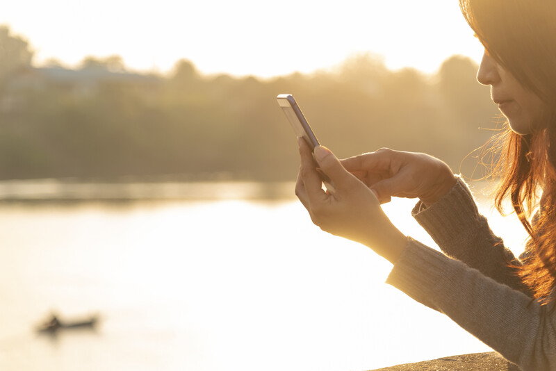 Woman holding smartphone by water at golden hour, backlighting creates lens flare effect.
