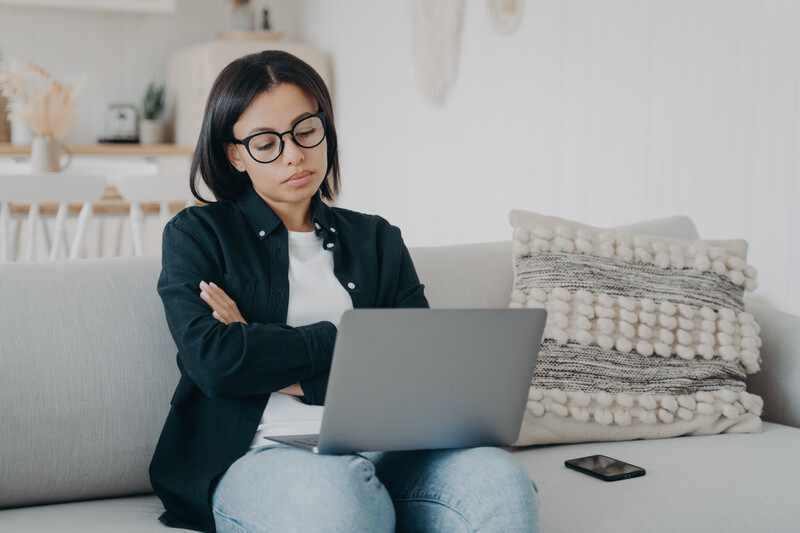 Woman with glasses working on laptop while sitting on couch with arms crossed.