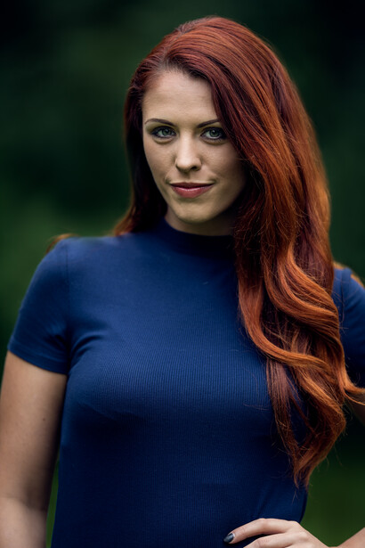 Woman with long reddish-brown hair wearing a dark blue short-sleeve shirt against a soft green blurred background.