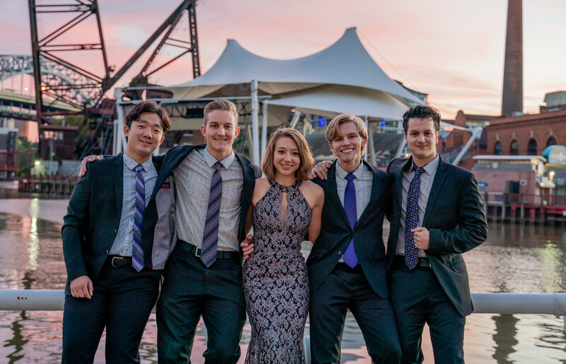 Five people in business formal attire standing together at an urban waterfront during golden hour.