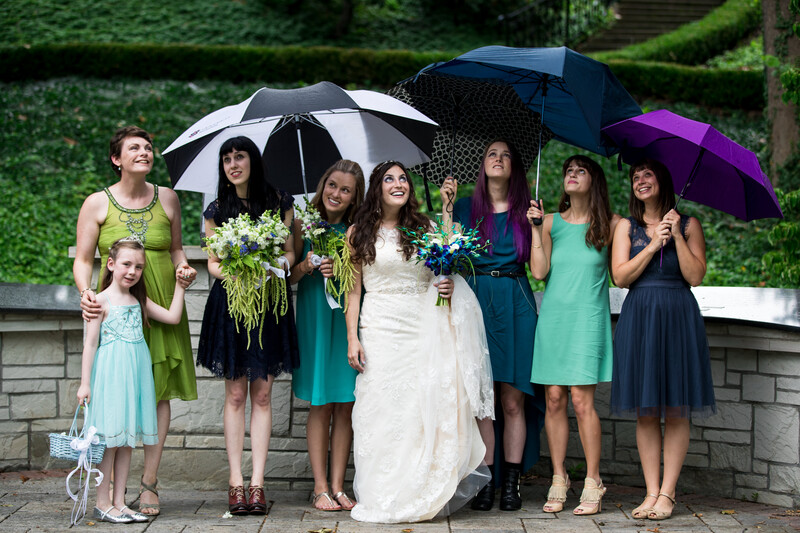 Bride in white dress with wedding party holding umbrellas in rain outdoors.