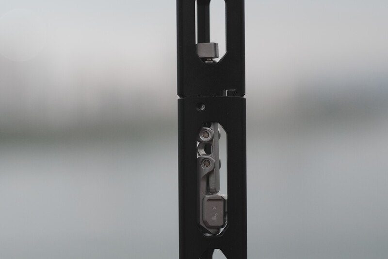 Close-up of a black vertical rail or pole with mounted mechanical hardware and metal fittings against a blurred gray background.