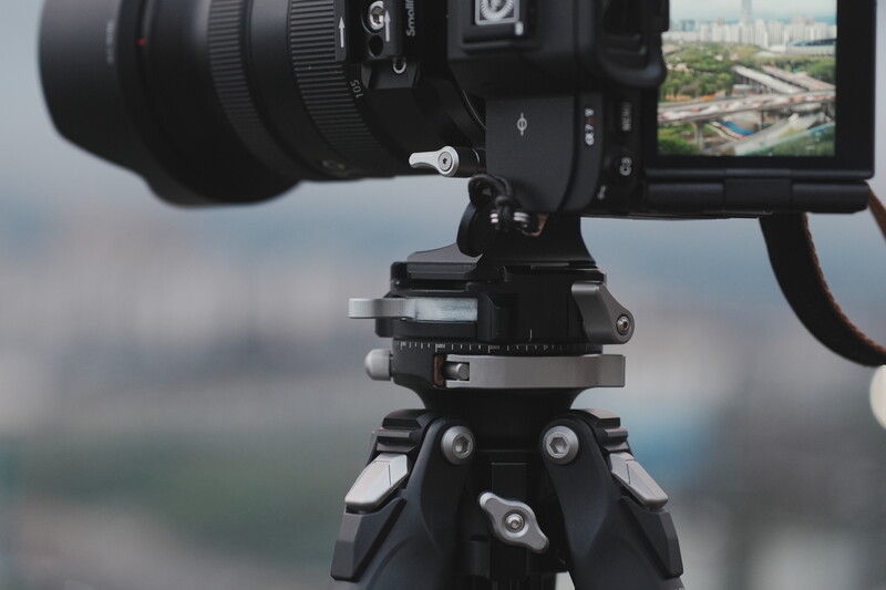 Professional video camera mounted on a fluid head tripod with articulating arm, overlooking an aerial landscape view on the LCD monitor.