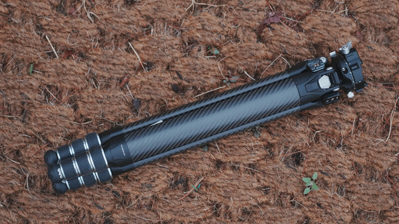 Carbon fiber camera tripod head with black metal components and adjustment rings on brown mulch background.