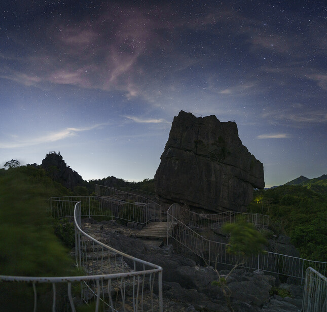 Metal viewing platform with railings winding around a dramatic dark rock formation against a blue sky.