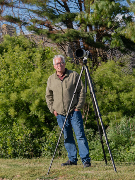 Photographer standing with camera mounted on tall tripod in outdoor setting with green foliage behind.