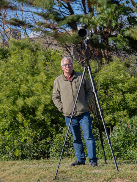 Photographer standing with camera mounted on tripod in outdoor setting with trees.