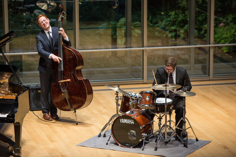 Cellist and drummer perform together in a modern concert hall with large windows overlooking trees.