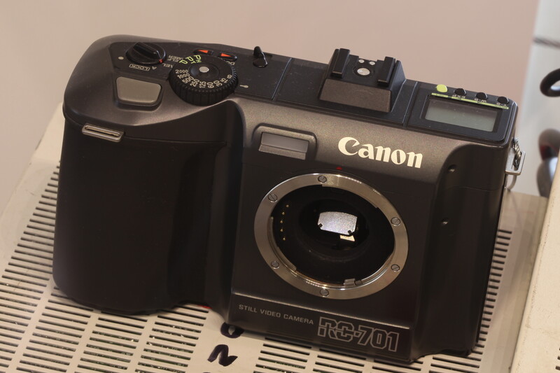 Canon RC-701 digital camera body without lens, showing open shutter and top-mounted controls against a white textured surface.