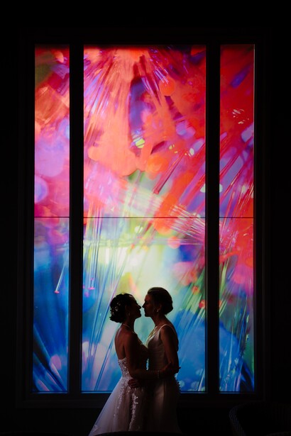 Silhouetted couple standing before a large triptych window displaying vibrant abstract light patterns in reds, purples, blues, and yellows.