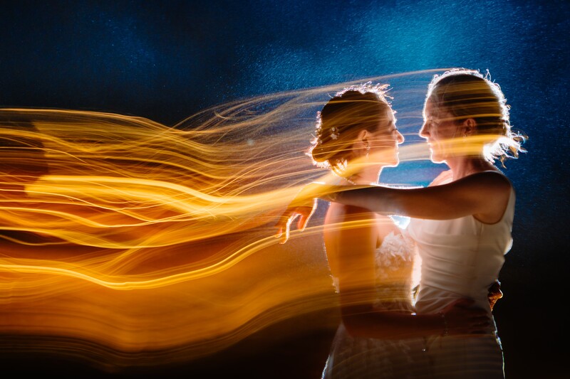 Two women in white tank tops with long golden light trails flowing from their bodies against a dark blue background.