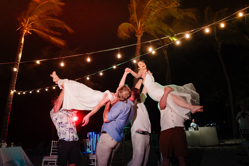 Group of people lifting a bride in white dress at outdoor evening reception with string lights and palm trees.