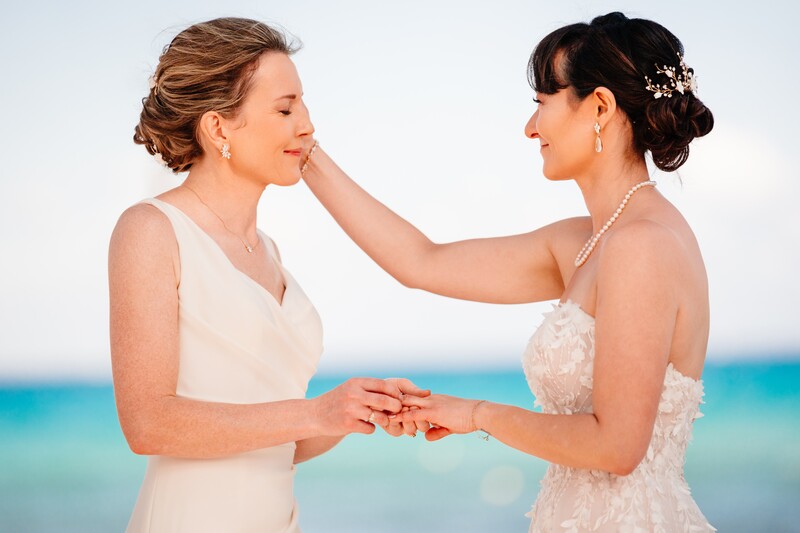 Two women in white dresses exchanging rings on a beach with turquoise ocean behind them.