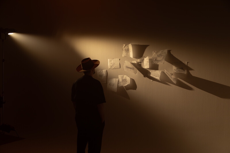 Silhouetted figure in hat standing before wall with projected shadow and geometric text shapes.