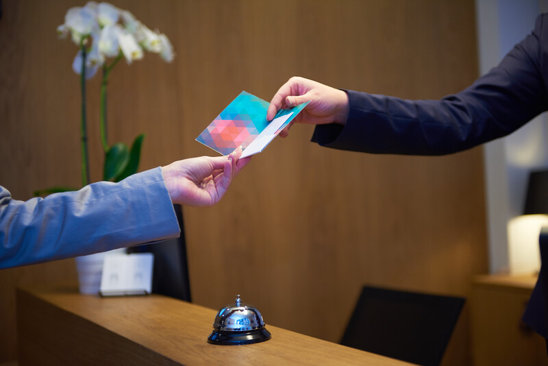 Two people exchanging a colorful card or print at a wooden reception desk with a service bell and flowers.
