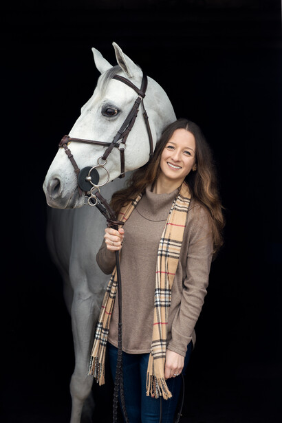 Woman in brown cardigan holding a gray horse's bridle against a black background.