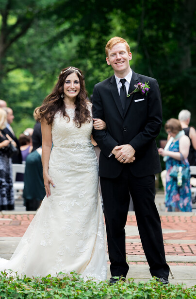 Bride and groom walking together outdoors during wedding ceremony with guests visible in background.