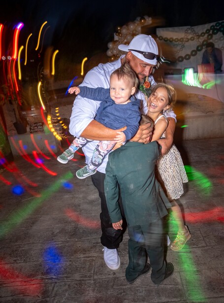 Adult holding two children while standing on a colorful lit dance floor at night.