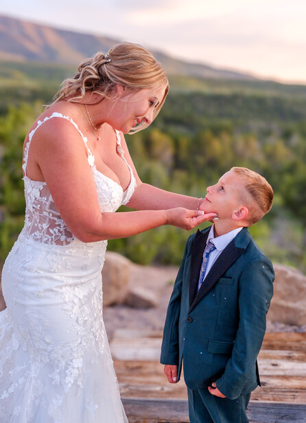 Bride in white lace dress holding hands with young boy in navy suit outdoors.