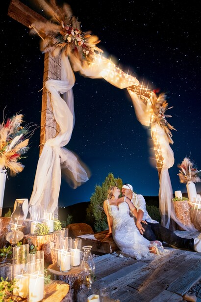 Couple kissing under an illuminated fabric arch at a nighttime outdoor wedding ceremony beneath starry sky.
