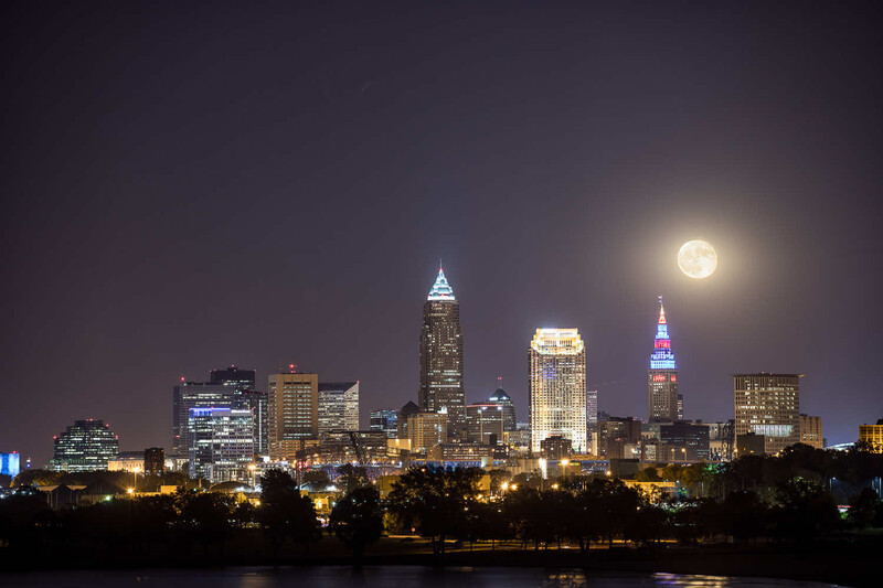 Full moon rising above a cityscape with illuminated skyscrapers reflected in water below.