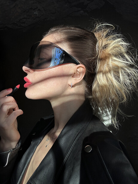 Woman wearing dark sunglasses and black blazer with blonde hair in bun against dark background.