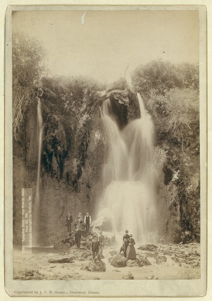 Tall waterfall cascading down rocky cliff surrounded by dense vegetation and trees.