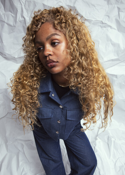 Portrait of a woman with long curly golden-blonde hair wearing a dark blue denim shirt against a white textured backdrop.