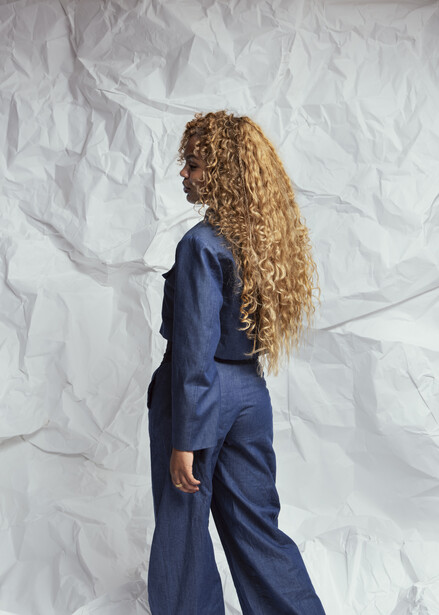 Woman with long curly blonde hair wearing a navy blue suit, photographed from behind against a white textured backdrop.