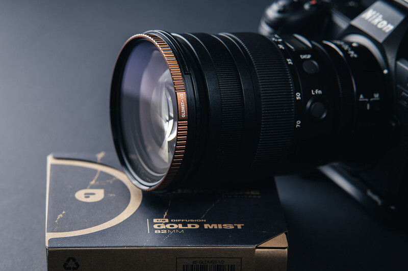 Black camera lens with copper-colored focus ring mounted on DSLR body, positioned above a lens box on gray surface.