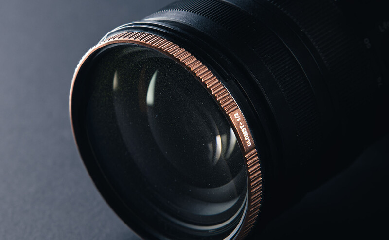 Close-up of a camera lens with copper-toned focusing ring and black glass elements against neutral background.