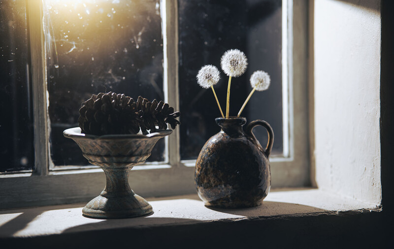 Still life arrangement with dandelion seedheads in a dark pitcher and a shallow dark vessel on a sunlit windowsill.