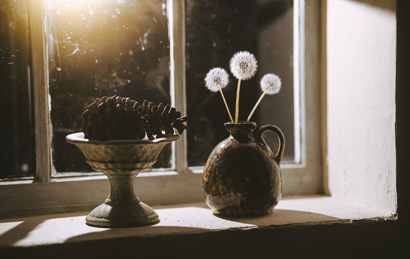 Dandelion seedheads in a weathered brass pitcher beside a dark vessel on a sunlit windowsill.