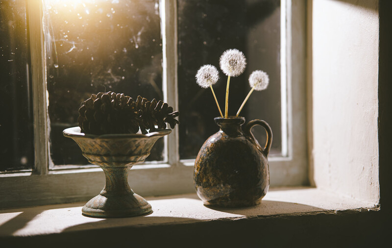 Two vintage vessels on a windowsill with dandelion seedheads backlit by golden sunlight streaming through glass panes.