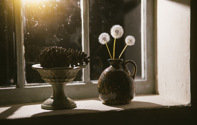 Three dandelion seedheads in a ceramic pitcher on a sunlit windowsill beside a decorative urn.