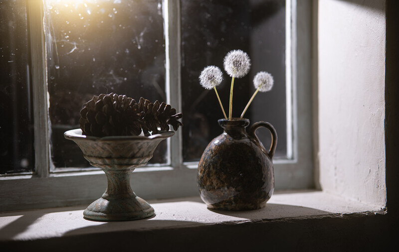 Still life with dandelion seedheads in a dark ceramic pitcher and a textured bowl on a sunlit windowsill.