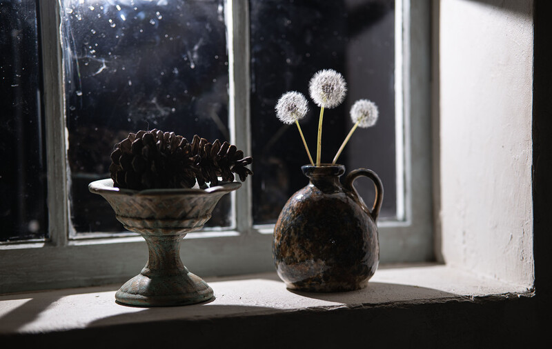 Weathered ceramic vessels on a sunlit windowsill: a dark bowl with spiky seedpod and a pitcher holding three delicate dandelion seed heads.